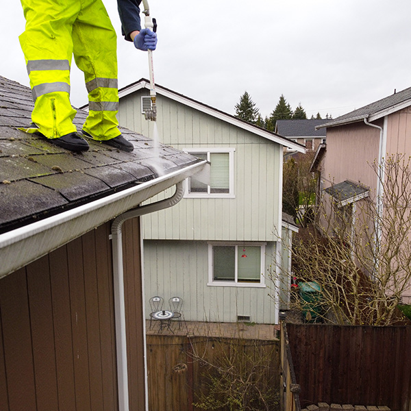 professional in yellow work pants standing on roof of brown home cleaning it